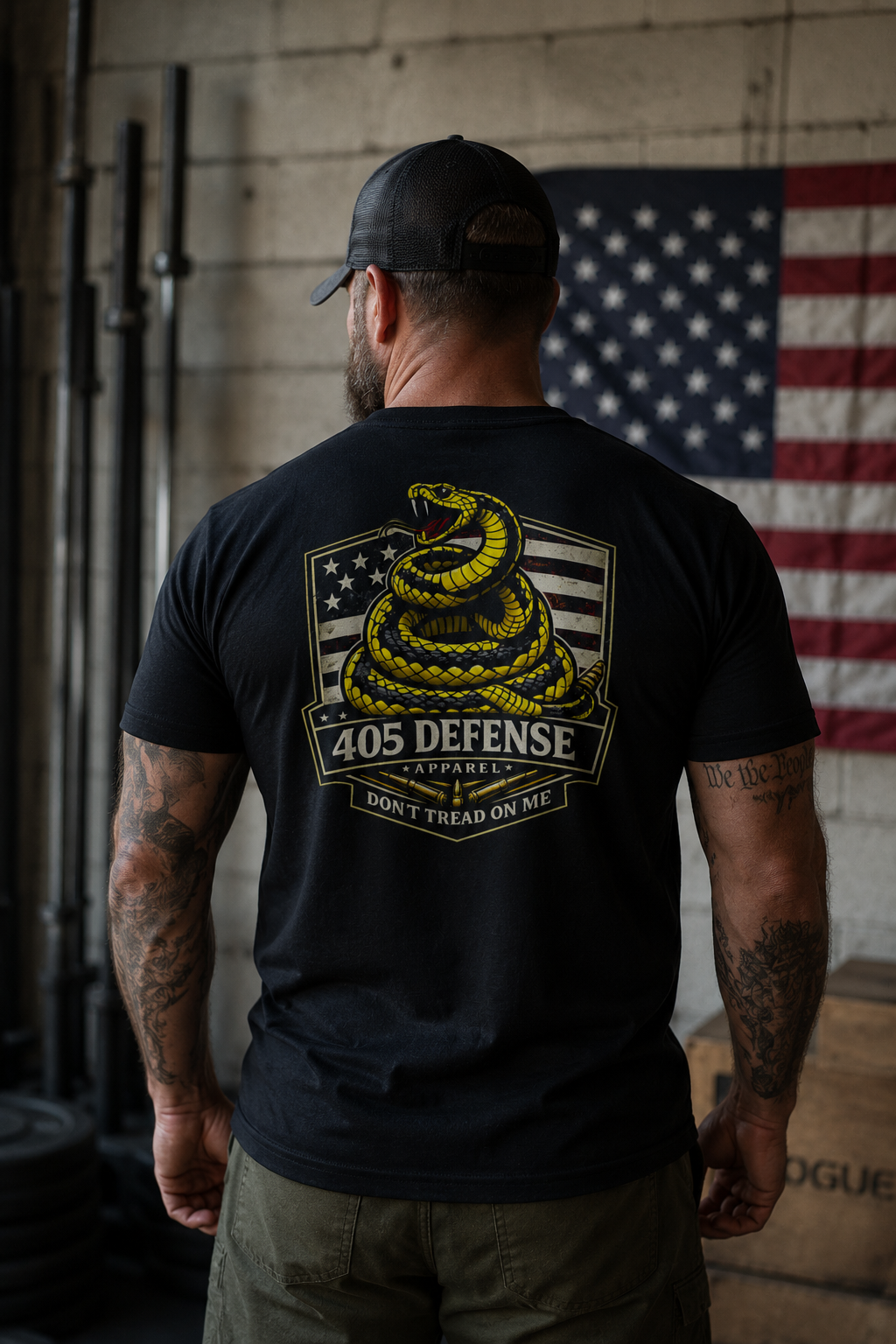 Back view of a muscular man wearing a black 405 Defense Apparel T-shirt featuring a large yellow coiled snake and distressed American flag graphic with “405 Defense Apparel” and “Don’t Tread on Me” text, standing in an industrial gym with tattooed arms, olive cargo pants, and an American flag in the background.