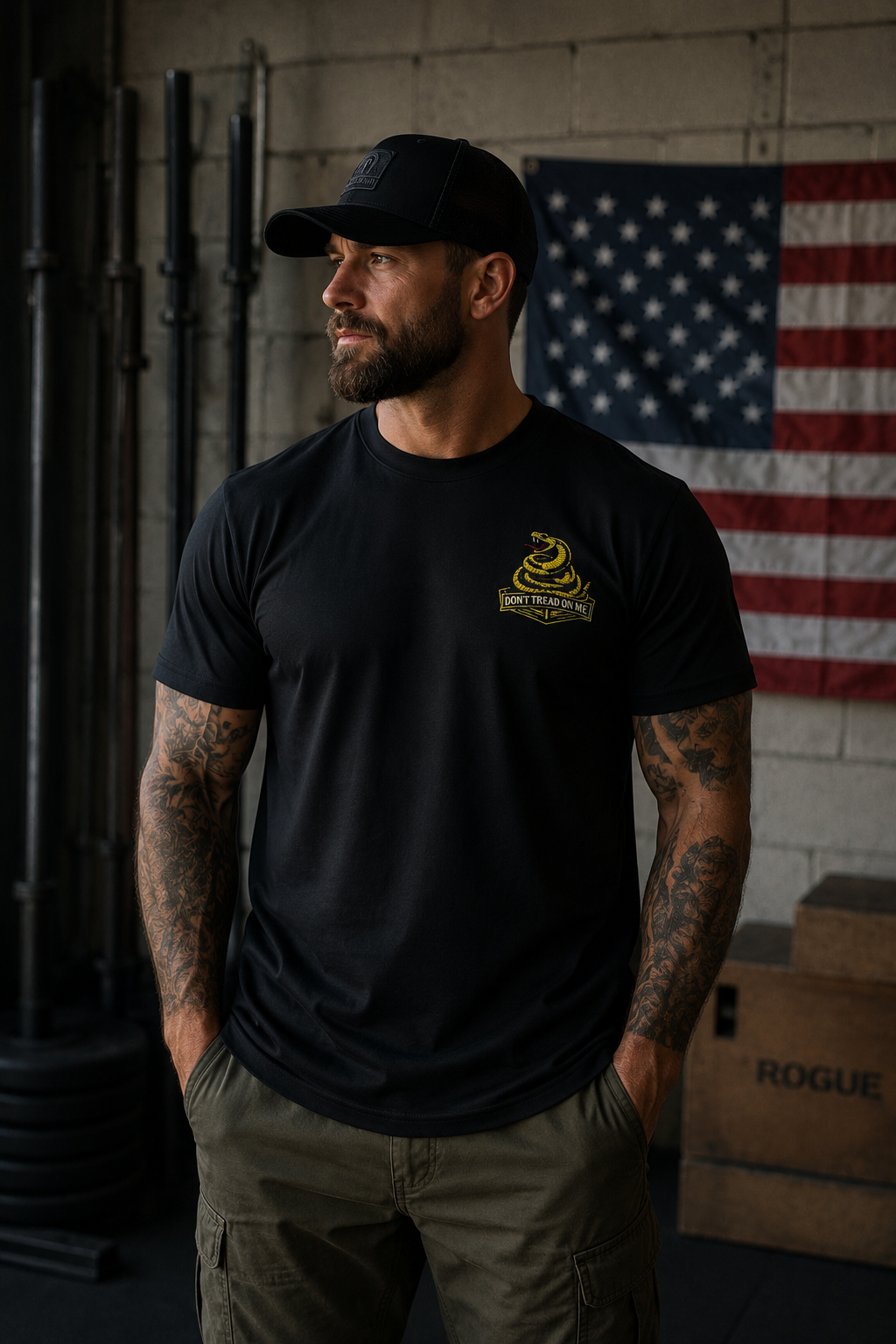 Muscular bearded man wearing a black 405 Defense Apparel T-shirt with a yellow “Don’t Tread on Me” snake chest logo, standing in an industrial-style gym with tattooed arms, olive cargo pants, a black trucker hat, and an American flag hanging in the background.
