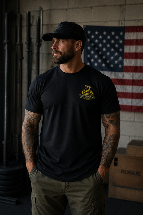 Muscular bearded man wearing a black 405 Defense Apparel T-shirt with a yellow “Don’t Tread on Me” snake chest logo, standing in an industrial-style gym with tattooed arms, olive cargo pants, a black trucker hat, and an American flag hanging in the background.