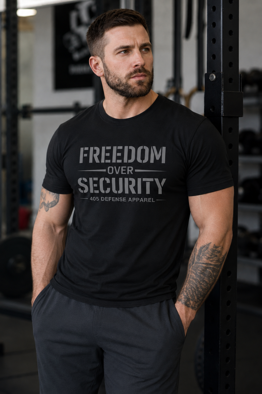 Athletic man wearing a black “Freedom Over Security” graphic T-shirt while standing in a modern gym next to a power rack. The shirt features a clean gray stencil-style print with “405 Defense Apparel” underneath, paired with dark athletic pants and visible forearm tattoos.
