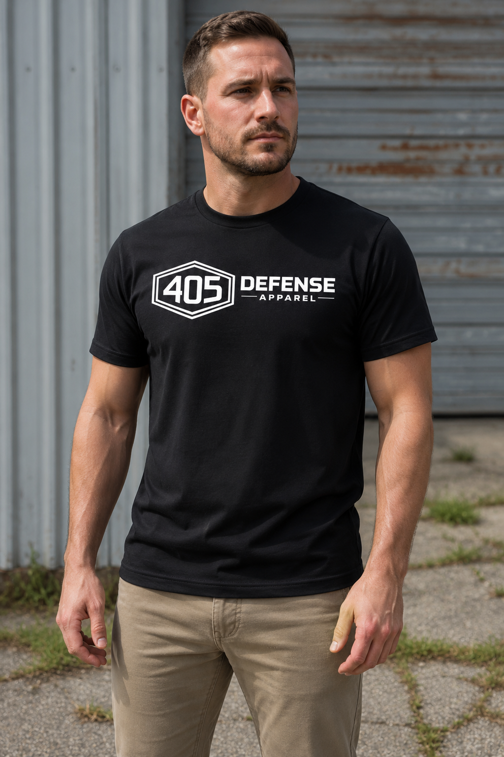 Athletic man wearing a black 405 Defense Apparel t-shirt with a bold white chest logo, standing outdoors in an industrial setting with metal siding and a warehouse-style background. The model is wearing khaki pants and looking off to the side in natural daylight, showcasing the front design in a realistic lifestyle apparel photo.