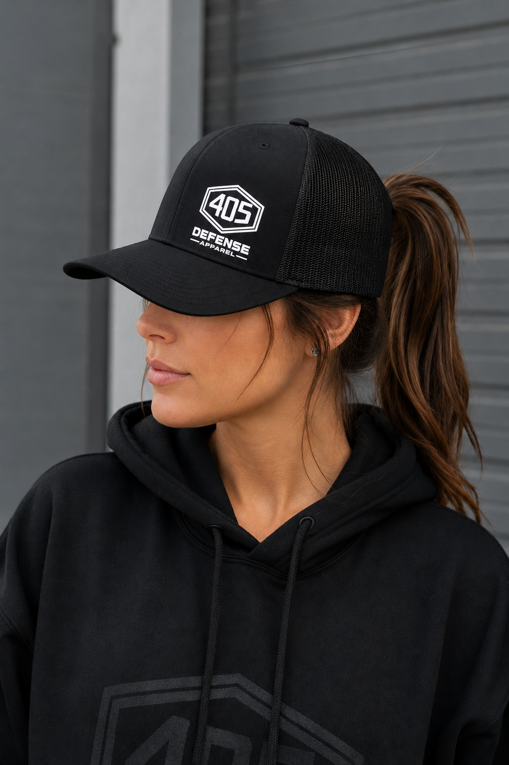 Female model wearing a black 405 Defense Apparel trucker hat with white logo embroidery and a black mesh back, shown in a realistic outdoor lifestyle setting with a ponytail pulled through the back opening of the cap.