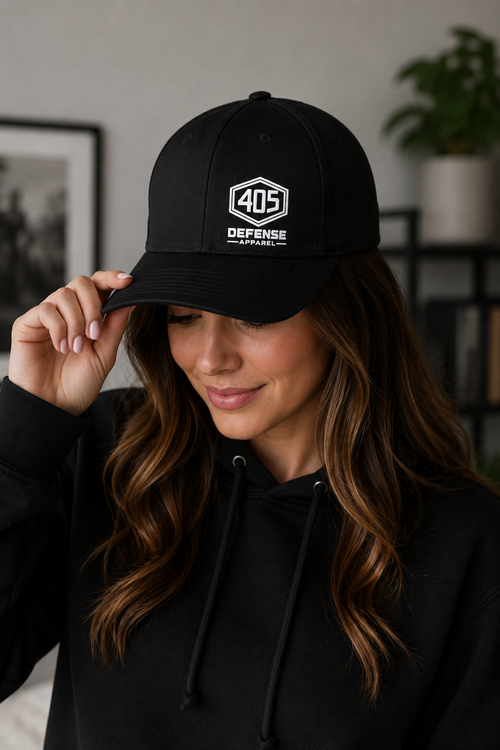 Woman wearing a black Flexfit 405 Defense Apparel hat with white hex logo embroidery, holding the brim of the hat while wearing a black hoodie in a softly lit indoor lifestyle setting.