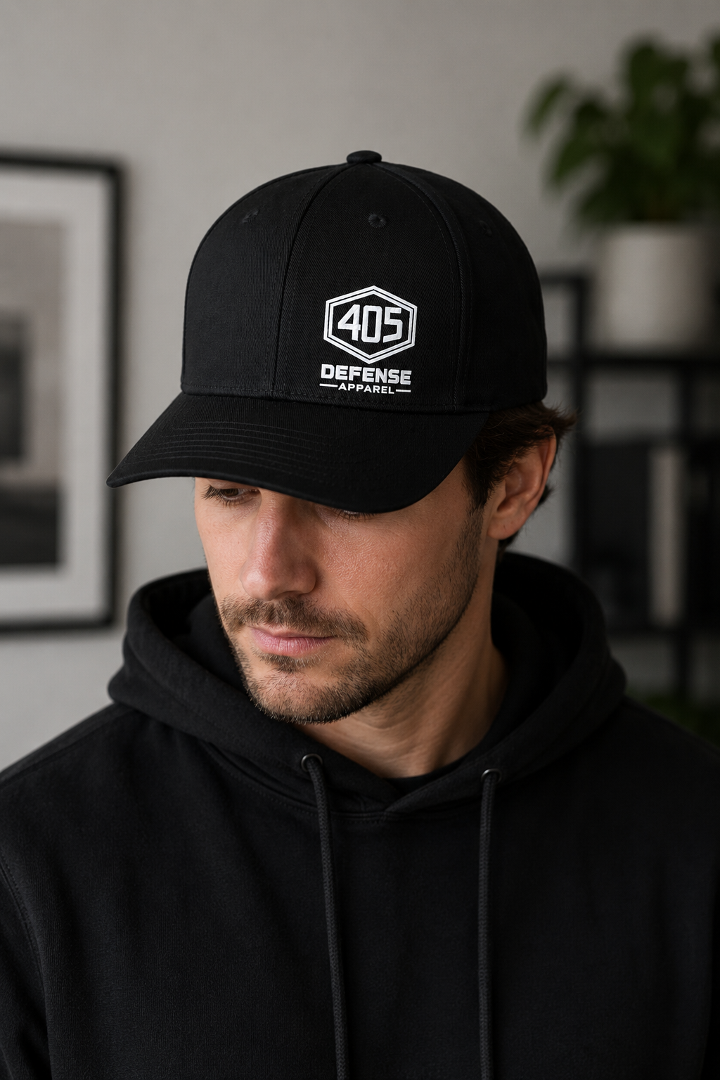 Man wearing a black Flexfit 405 Defense Apparel hat with white hex logo embroidery, paired with a black hoodie in a modern indoor setting with soft natural lighting.