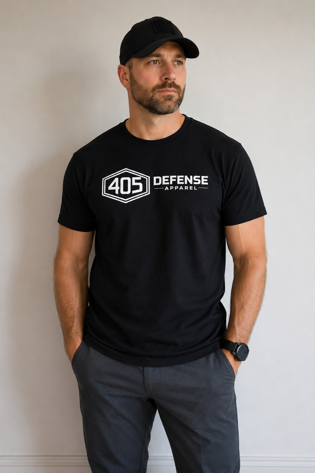 Man wearing a black 405 Defense Apparel t-shirt and matching black Flexfit hat with white logo branding, standing against a light gray studio background with hands in pockets.