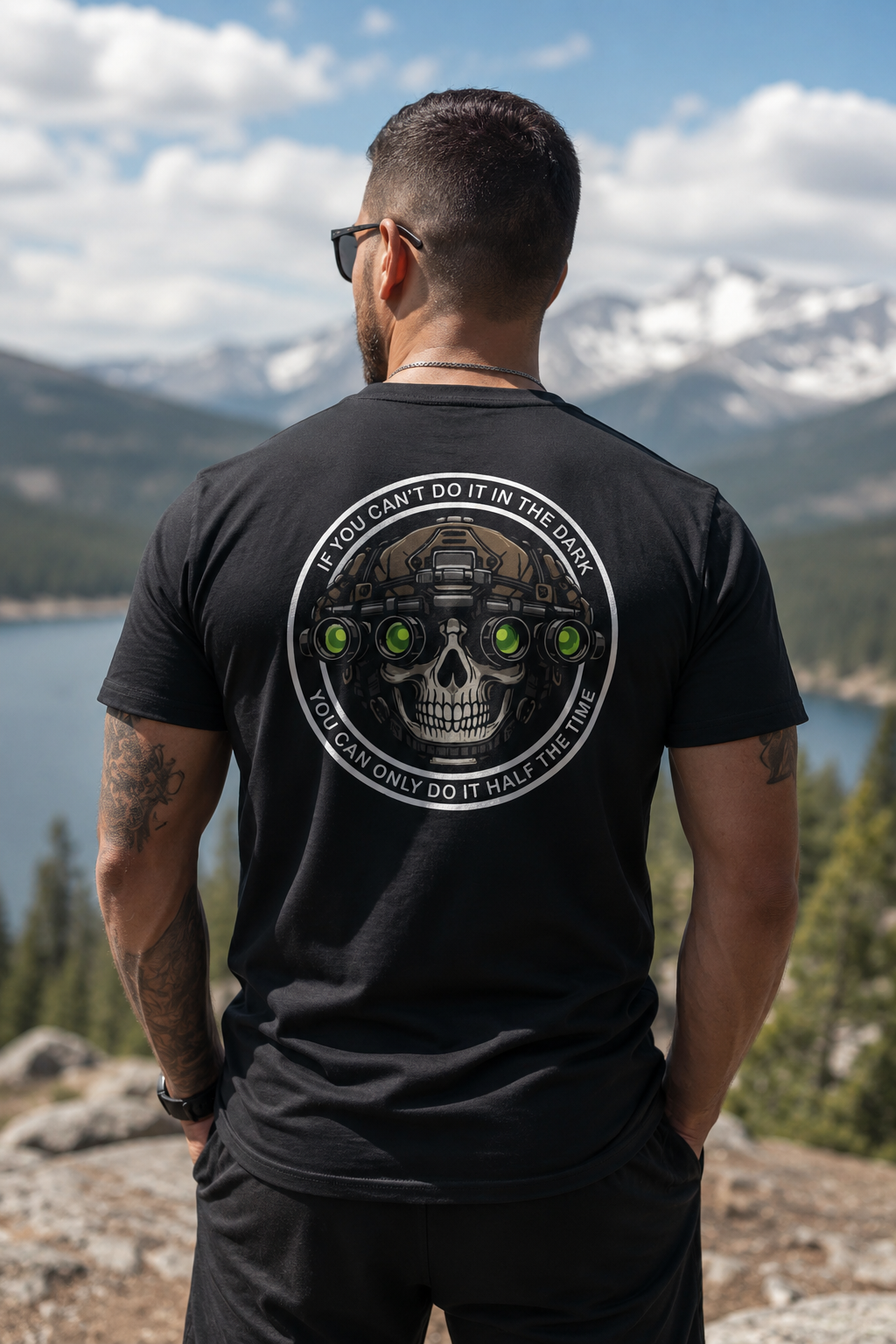 Man standing outdoors with his back facing the camera, wearing a black tactical-style T-shirt featuring a large skull graphic with night vision goggles and the phrase “If You Can’t Do It In The Dark You Can Only Do It Half The Time.” Mountain and lake scenery blurred in the background under a bright sky.
