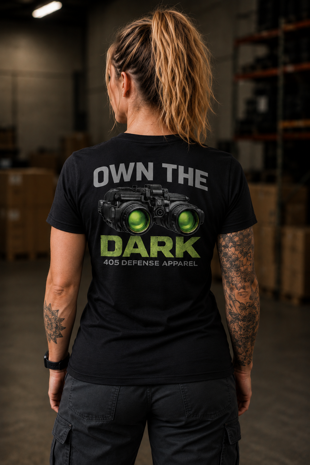 Back view of a tattooed woman standing in an industrial warehouse wearing a black “Own The Dark” 405 Defense Apparel t-shirt featuring green night vision goggles graphic and bold gray and green lettering, paired with dark cargo pants under warm cinematic lighting.