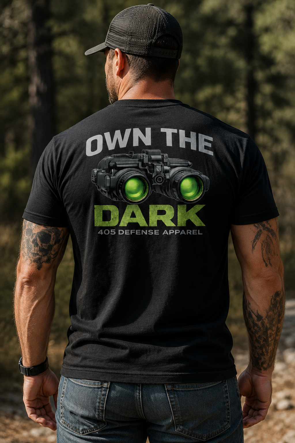 Muscular man standing with his back to the camera on a wooded trail wearing a black “Own The Dark” 405 Defense Apparel t-shirt featuring green night vision goggles graphic and bold distressed lettering, paired with jeans and a black mesh hat in warm outdoor lighting.