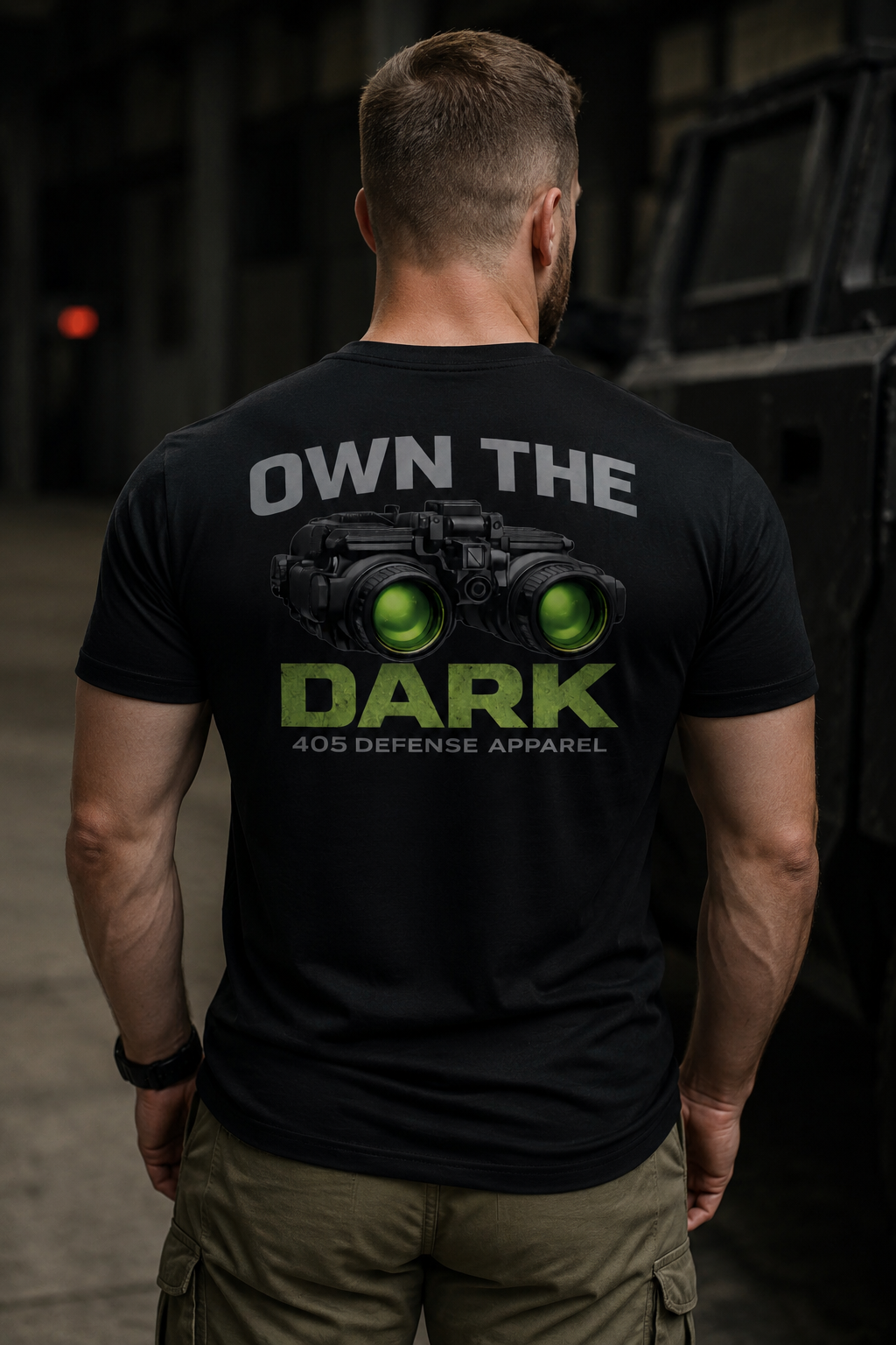Back view of a muscular man wearing a black “Own The Dark” 405 Defense Apparel t-shirt with glowing green night vision goggles graphic, standing inside a dark industrial warehouse with cinematic moody lighting and tactical-style cargo pants.