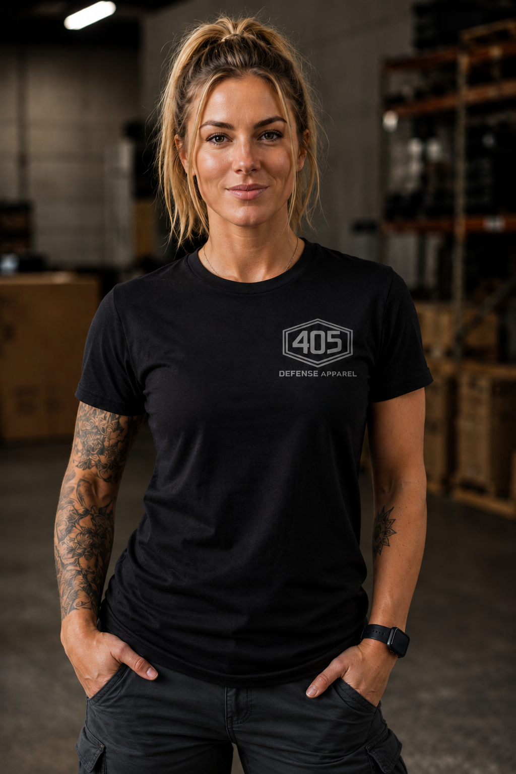 Confident woman standing in an industrial warehouse wearing a black 405 Defense Apparel t-shirt with a gray hex logo chest print, paired with dark cargo pants and tattoos, photographed in warm cinematic lighting with a blurred warehouse background.