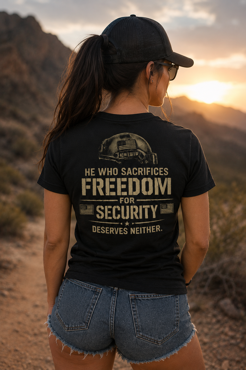 Rear view of a female model wearing a black graphic t-shirt featuring a distressed tan tactical helmet design and the quote “He Who Sacrifices Freedom for Security Deserves Neither.” The woman is standing on a desert mountain trail at sunset wearing denim shorts, a black trucker hat, and sunglasses with golden sunlight and rocky terrain in the background.