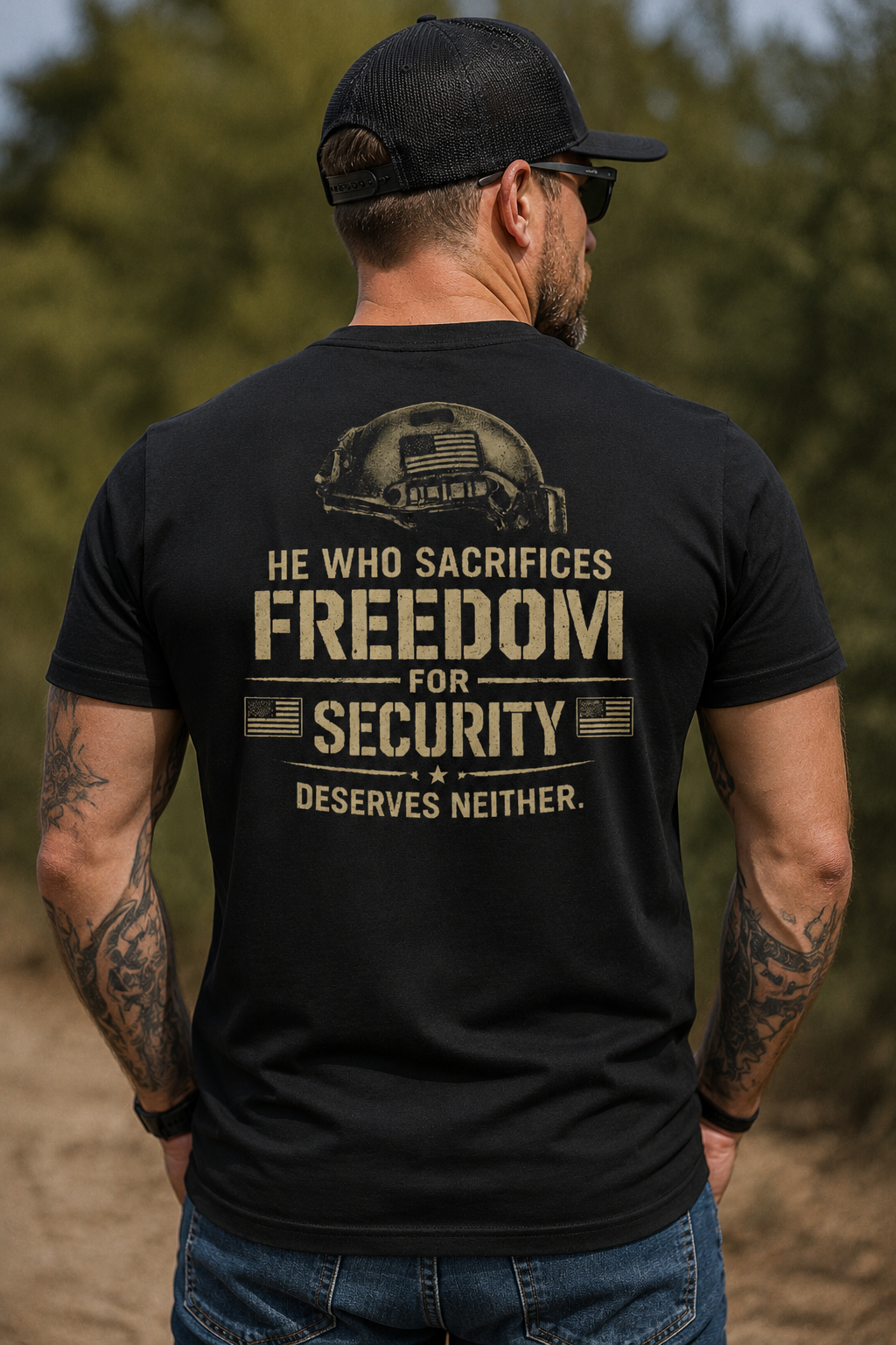 Rear view of a tattooed man wearing a black t-shirt with a large distressed tan graphic featuring a tactical helmet and the quote “He Who Sacrifices Freedom for Security Deserves Neither.” The male model is standing outdoors on a dirt path wearing blue jeans, a black hat, and sunglasses with blurred green trees in the background.