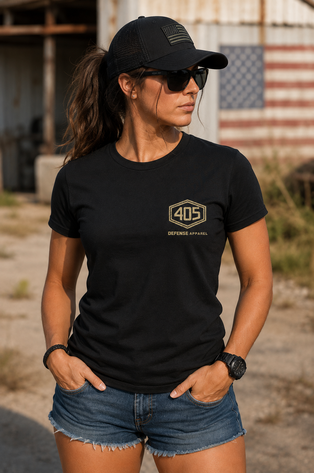 Female model wearing a fitted black 405 Defense Apparel t-shirt with a tan hex-style “405” chest logo, paired with denim shorts, black sunglasses, and a black tactical trucker hat. The woman is standing outdoors near a rustic industrial building with warm natural lighting and blurred mountains in the background.