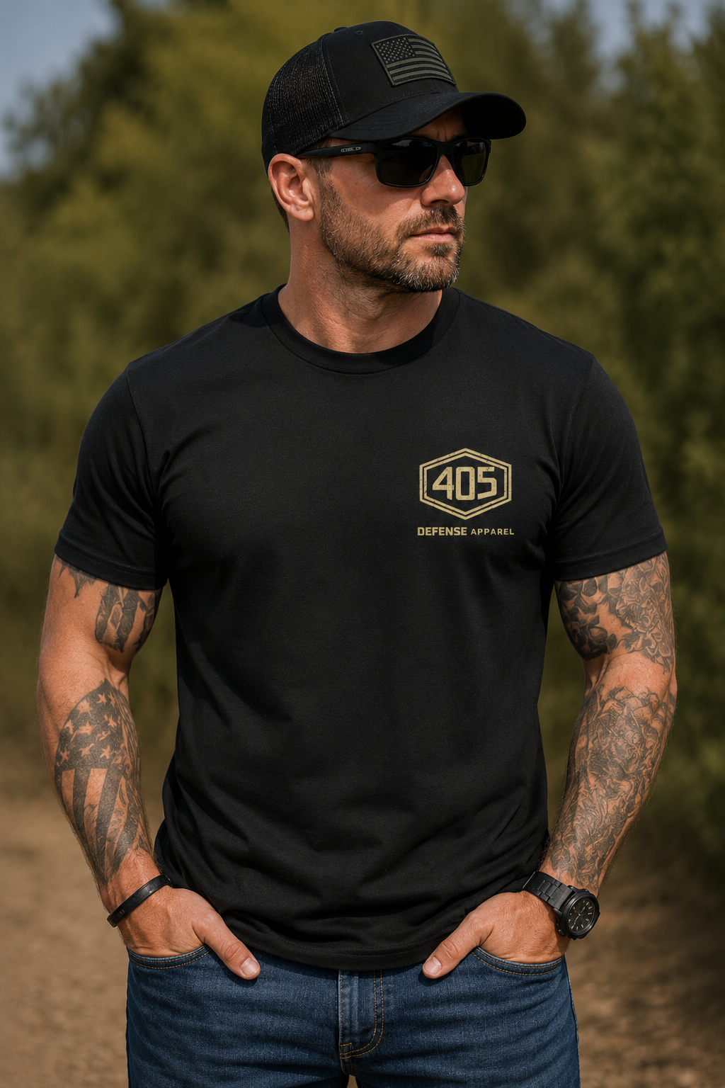 Muscular tattooed man wearing a black 405 Defense Apparel t-shirt outdoors, featuring a small tan hex-style “405” chest logo on the left side. The male model is wearing a black tactical-style hat, sunglasses, blue jeans, and a black watch while standing on a dirt trail with blurred trees in the background.
