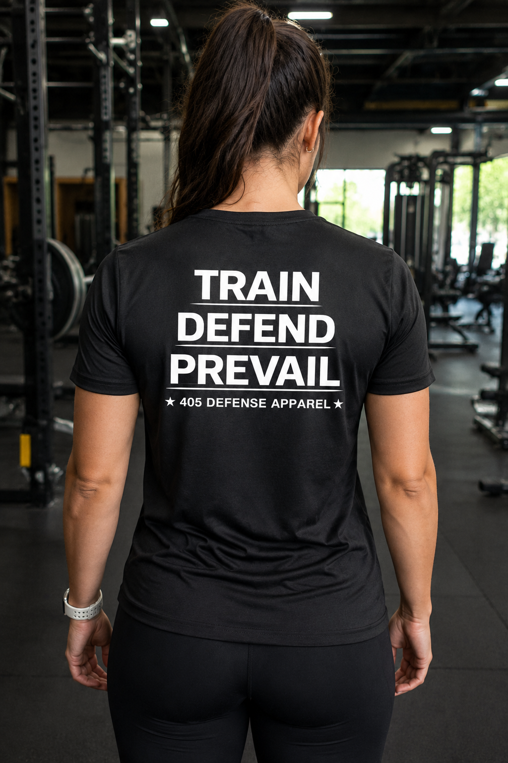 Female athlete viewed from behind in a gym wearing a black performance t-shirt featuring a large white “TRAIN DEFEND PREVAIL” graphic with “405 Defense Apparel” underneath. The athletic fit shirt is paired with black leggings, with squat racks and gym equipment visible in the background.