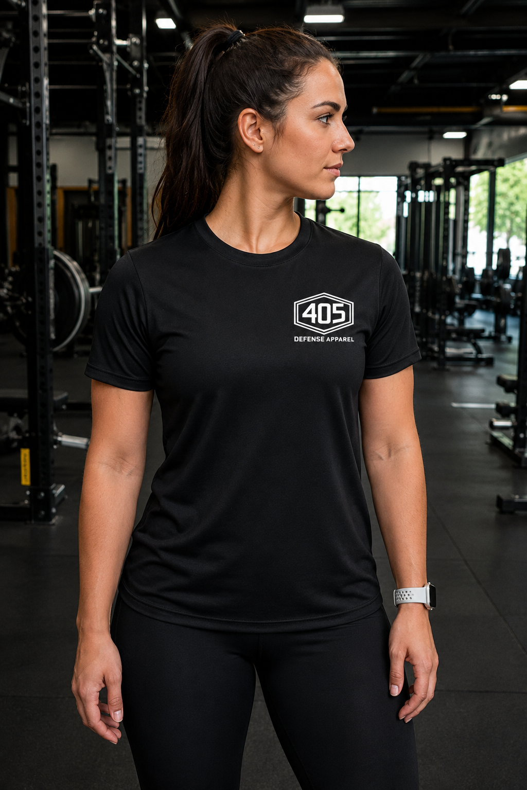 Female athlete standing in a modern gym wearing a black performance t-shirt with a white “405 Defense Apparel” hex logo on the left chest. The woman is shown from the front in black leggings, with gym equipment blurred in the background and industrial lighting creating a realistic fitness lifestyle scene.