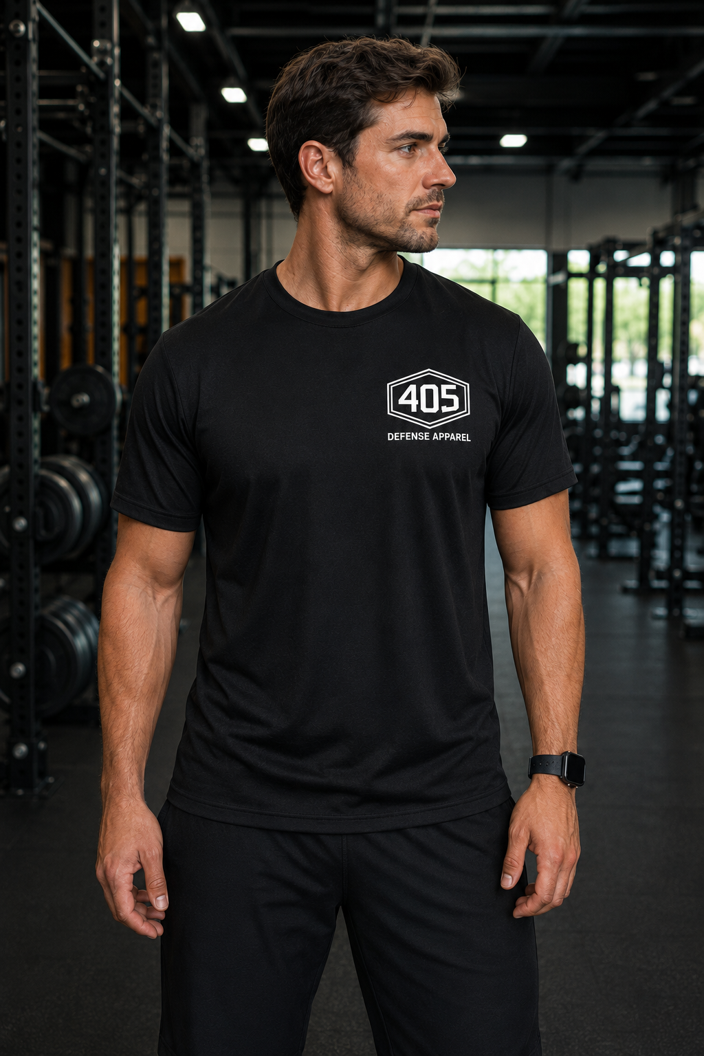 Male athlete standing in a modern gym wearing a black performance t-shirt with a white “405 Defense Apparel” hex logo on the left chest. He is shown from the front in black athletic shorts with gym equipment and squat racks blurred in the background under industrial lighting.
