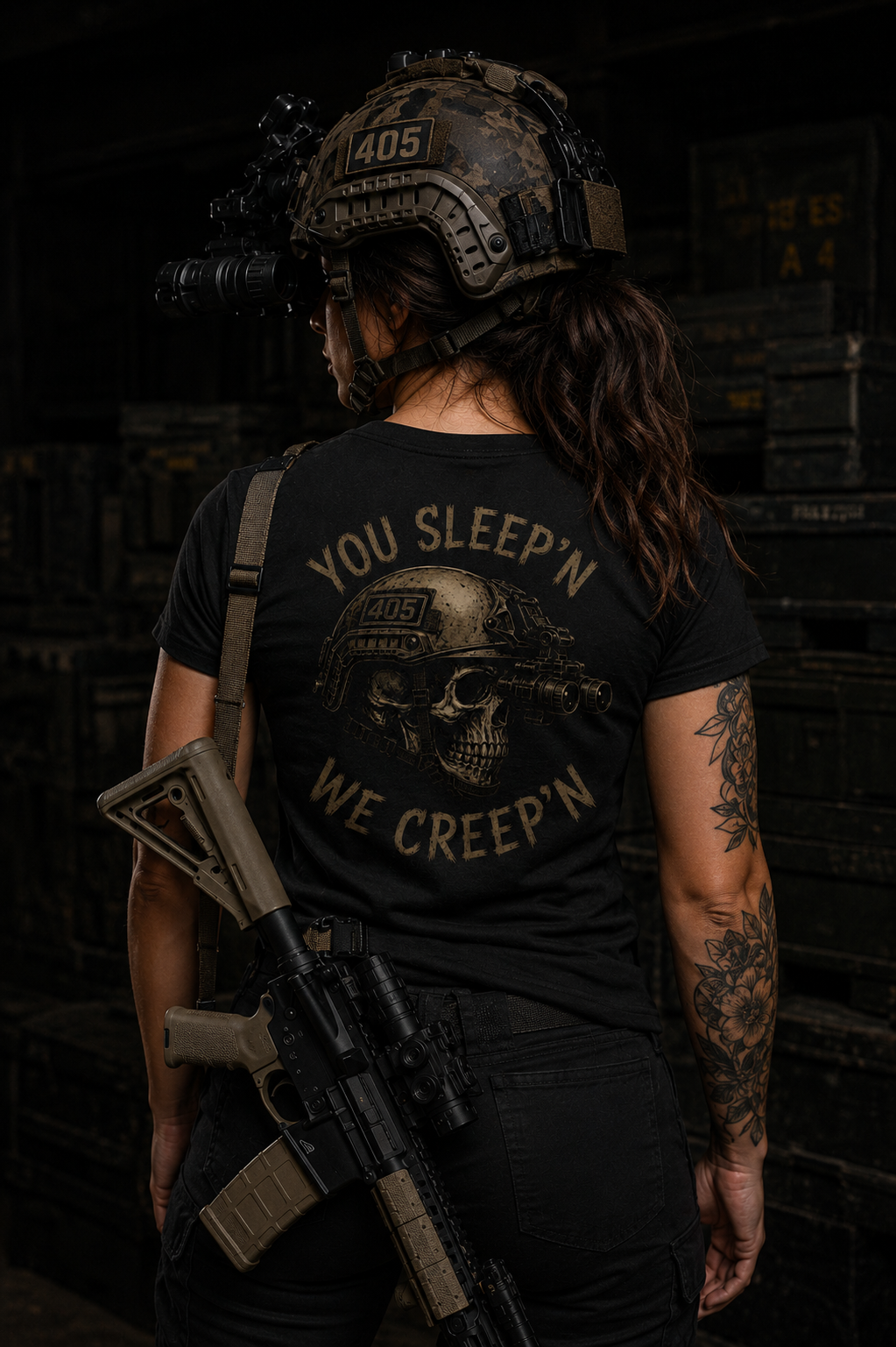 Female model standing in a dark industrial warehouse wearing a black tactical t-shirt with a distressed skull graphic and the phrase “YOU SLEEP’N WE CREEP’N” printed on the back in tan lettering. The model wears a tactical helmet with mounted night vision goggles and a “405” patch, with a rifle slung across her back. Dramatic low-light styling, tattooed arms, and military-inspired gear create a gritty tactical aesthetic.