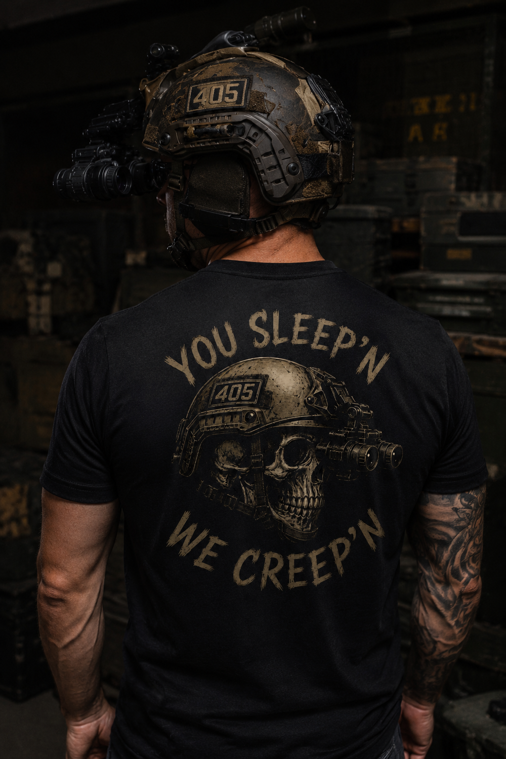 Muscular male model standing in a dark industrial warehouse wearing a black tactical-themed t-shirt with a large distressed skull graphic on the back. The design features a helmet with night vision goggles, a “405” patch, and the phrase “YOU SLEEP’N WE CREEP’N” in tan lettering. The model wears a tactical helmet with mounted night vision equipment and has tattooed arms, creating a gritty military-inspired aesthetic.