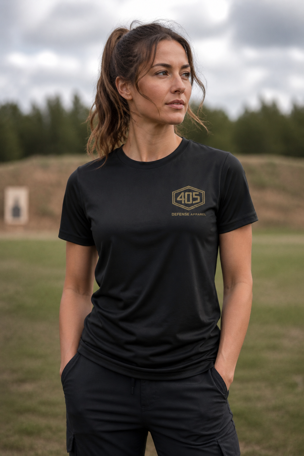 Female model standing outdoors at a shooting range wearing a black 405 Defense Apparel t-shirt with a tan hex logo chest print. The woman has her hands in her pockets and is looking off to the side, with a blurred grassy field and target in the background under an overcast sky.