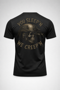 Back view of a black tactical graphic t-shirt featuring a skull wearing a helmet with night vision and the phrase “You Sleep’n We Creep’n” in distressed tan print.